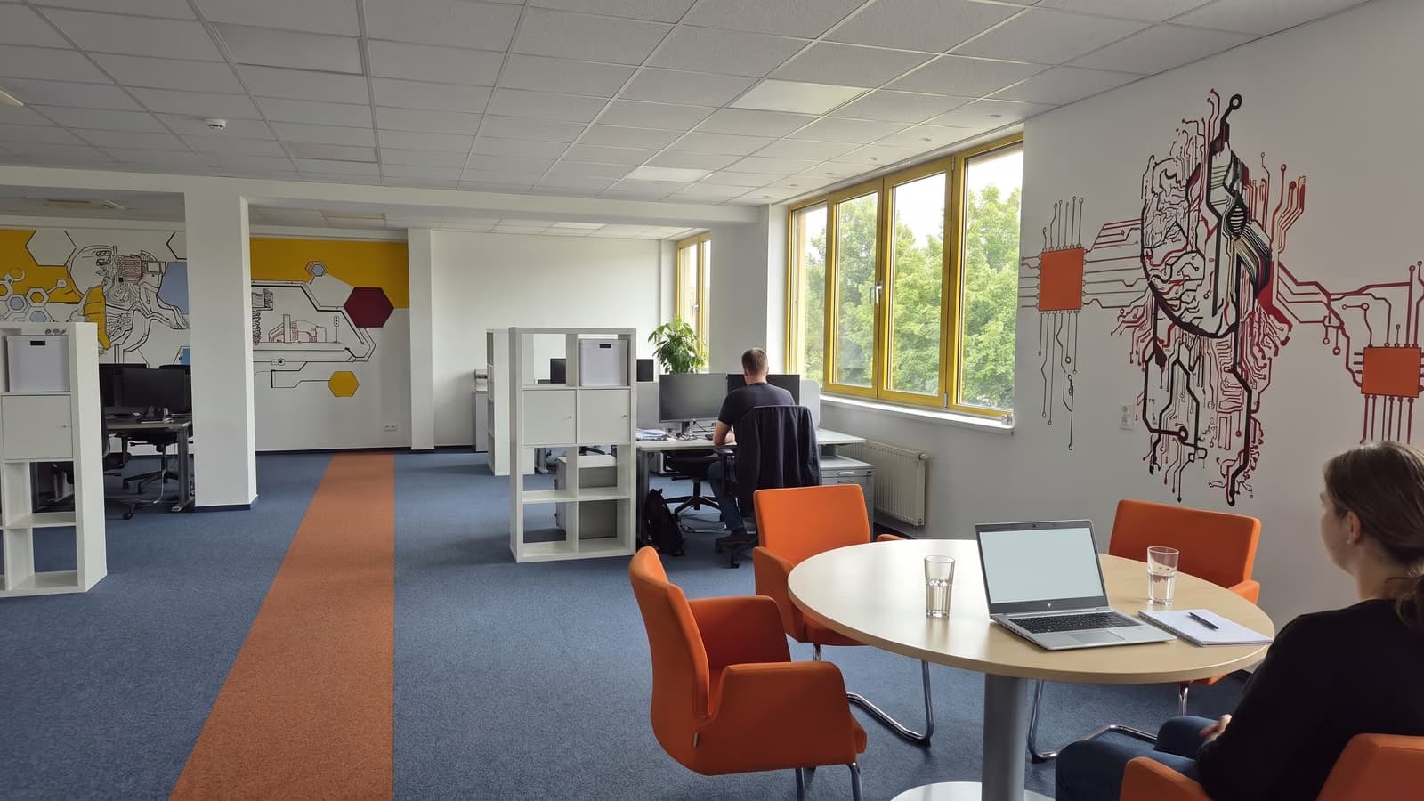 Beratungsgespräch an einem runden Tisch mit orangen Stühlen, ein aufgeklappter Laptop zwischen den Beteiligten, alle von hinten.