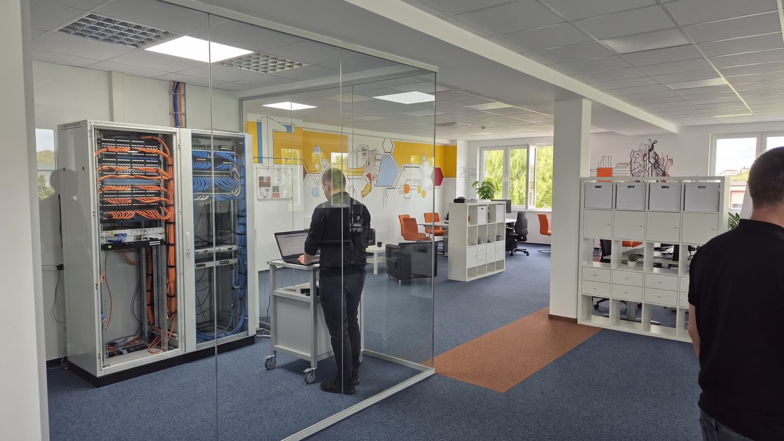 Blick vom Büro durch eine Glaswand in einen Technikraum mit zwei Netzwerkschränken, davor eine Person von hinten an einem Laptop auf einem Rollwagen.