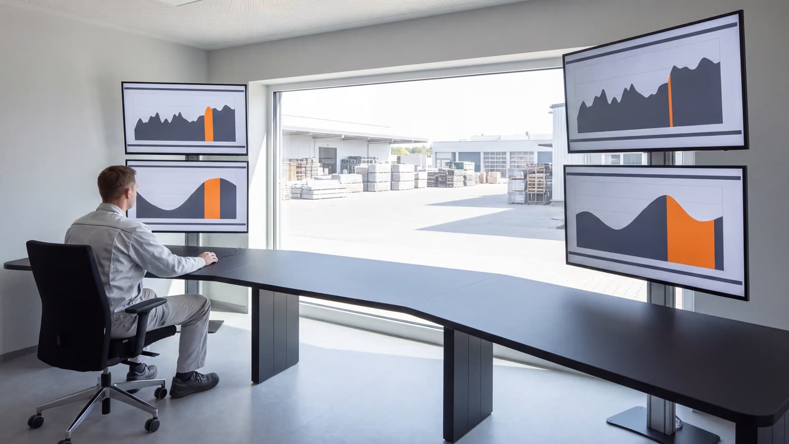 Leitstand mit vier Bildschirmen voller Verlaufskurven, eine Person davor, dahinter durch ein breites Fenster der Hof eines Industriegeländes bei Tageslicht.