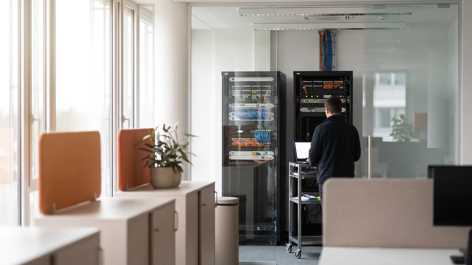 Blick vom Büro durch eine Glaswand in einen Technikraum mit zwei Netzwerkschränken, davor eine Person von hinten an einem Laptop auf einem Rollwagen.