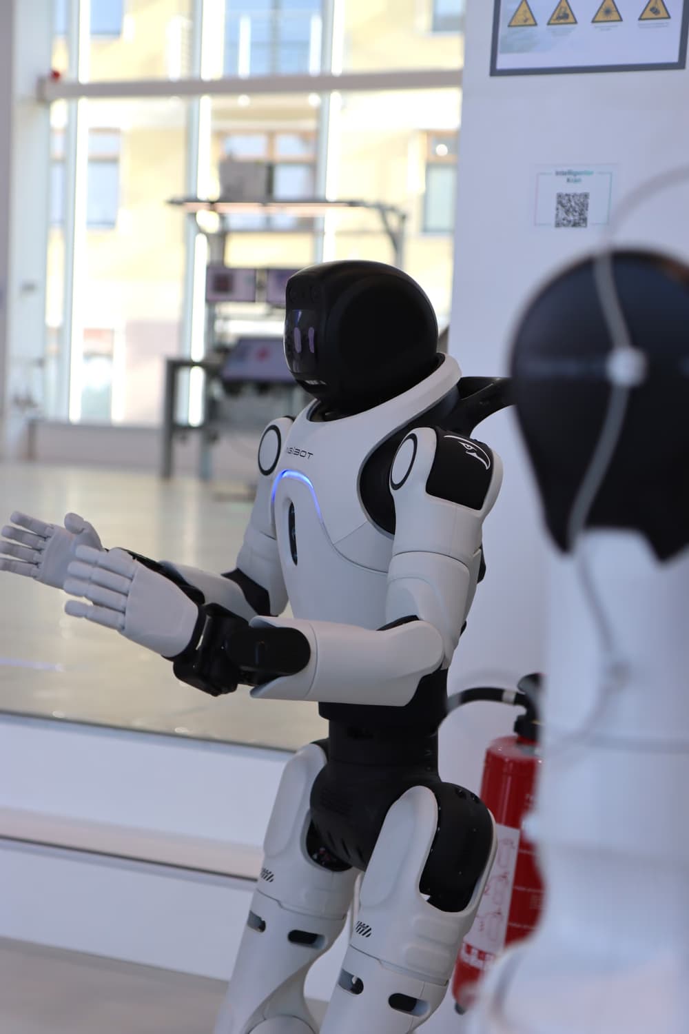 Ein humanoider Roboter in einer hellen Halle hebt beide Arme mit geöffneten Händen, hinter ihm steht ein zweiter Roboter.