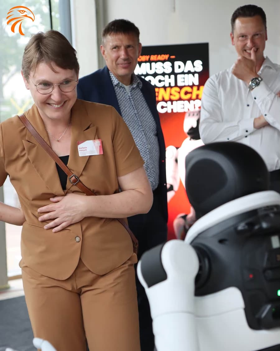 Drei Besucher der Eröffnung stehen lachend vor einem Roll-up mit der Frage, ob das noch ein Mensch machen muss; im Vordergrund ein humanoider Roboter.