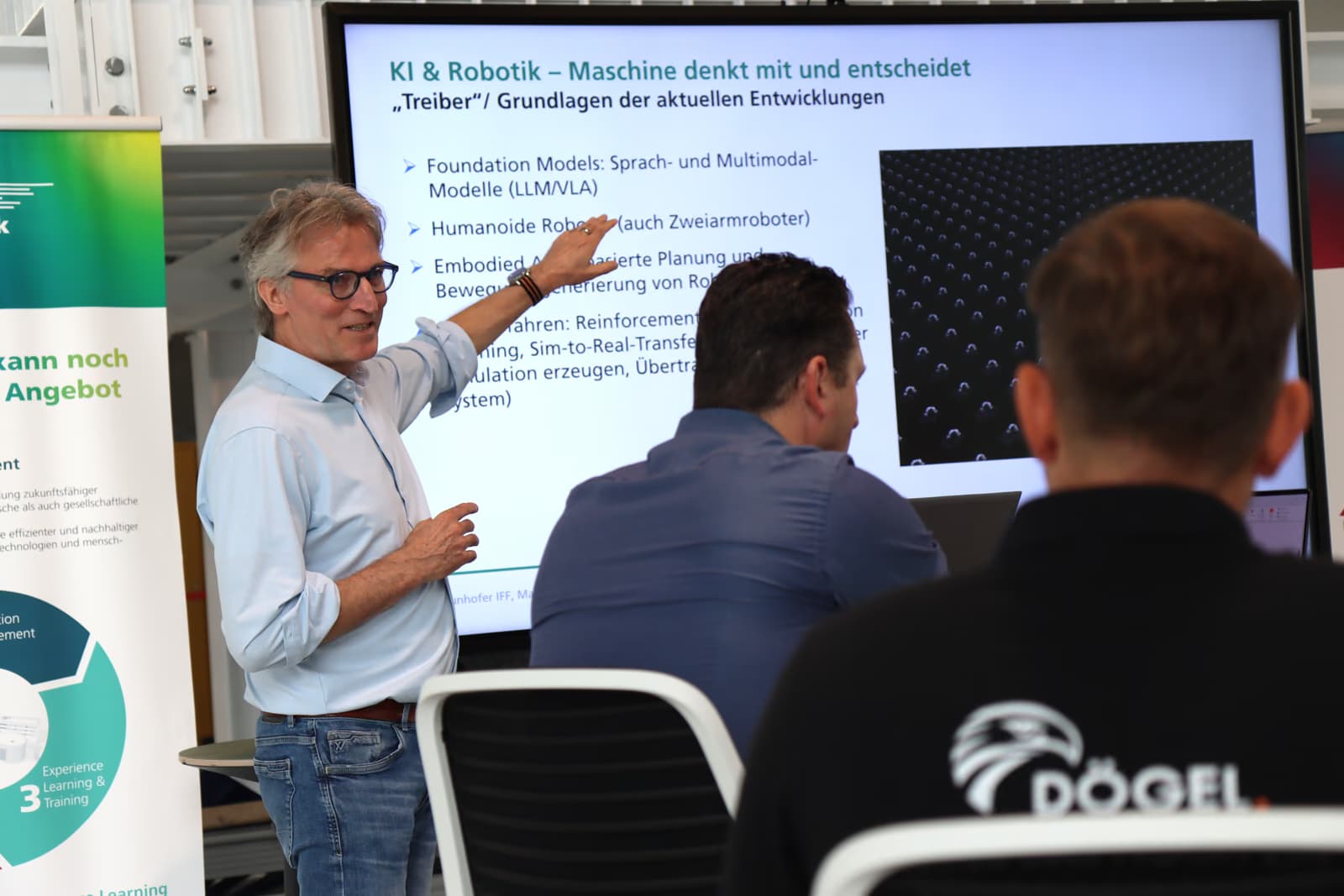 Ein Mann im hellen Hemd steht vor einer Gruppe an einem langen Tisch und zeigt auf einen Bildschirm mit einer Präsentation.