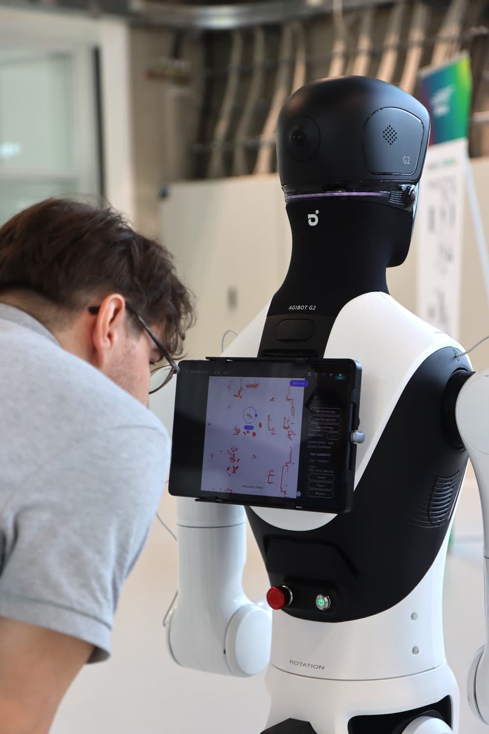 Ein humanoider Roboter von hinten, am Rücken ein Tablet mit einer Anzeige. Ein Mann mit Brille beugt sich heran und schaut auf den Bildschirm.