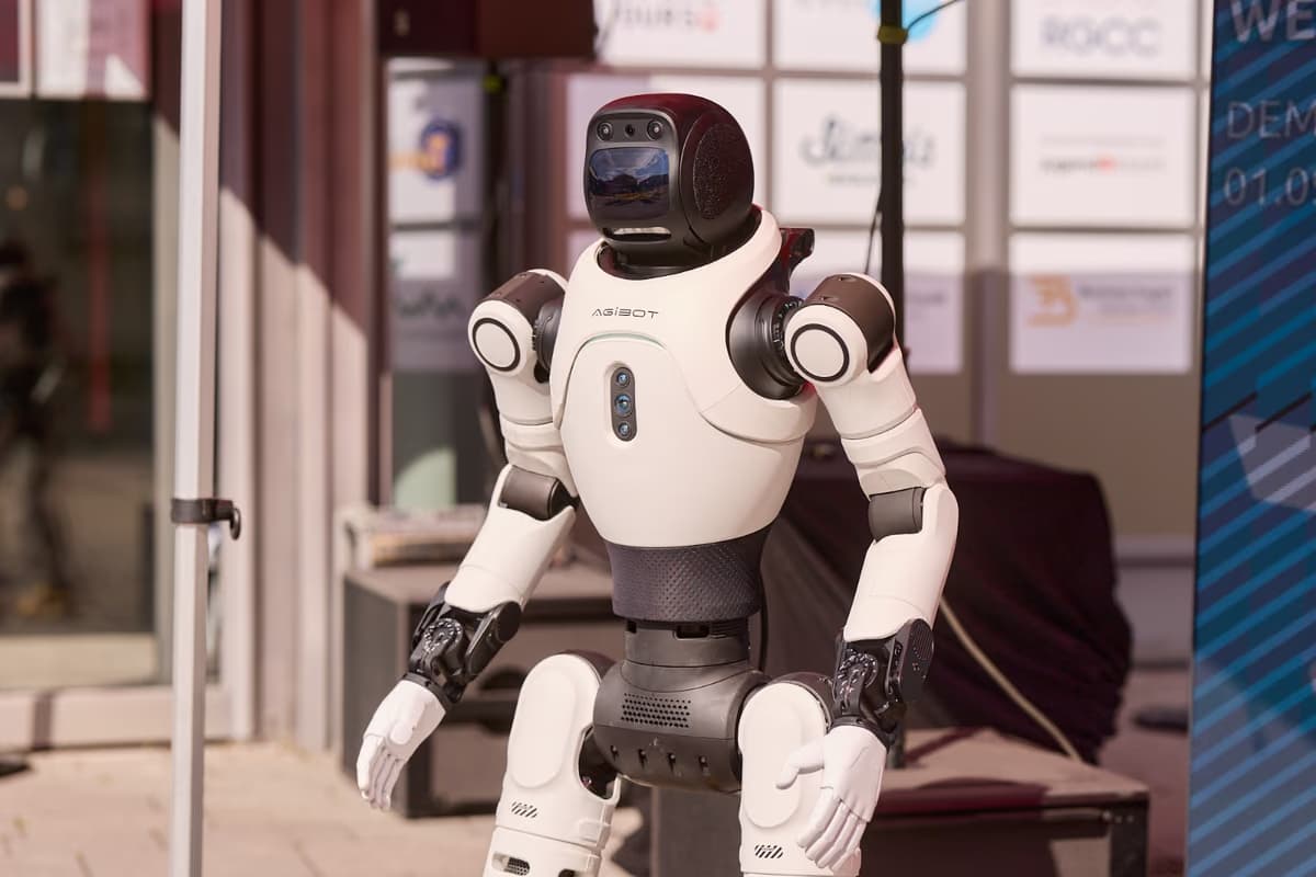 Nahaufnahme eines humanoiden Roboters mit weißer Schale und dunklen Gelenken, die Arme leicht angehoben, vor einer Fensterfront.