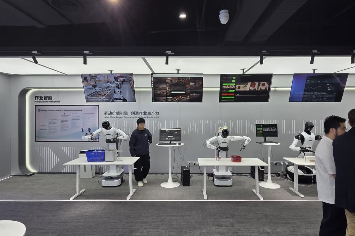 Mehrere Arbeitstische in einem Ausstellungsraum, an zwei von ihnen arbeiten humanoide Roboter, dazwischen stehen Besucher.