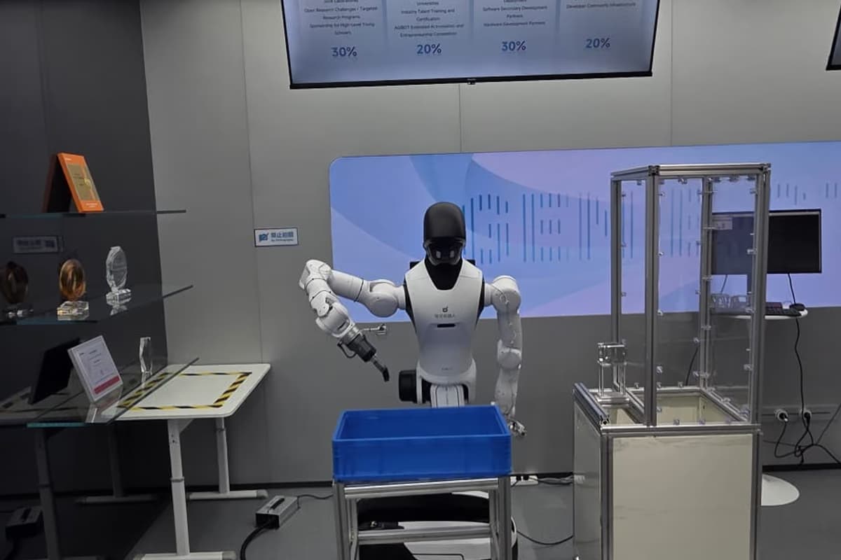 Nahaufnahme einer Arbeitsstation: ein humanoider Roboter hebt ein Teil aus einer blauen Kiste, dahinter ein Prüfaufbau aus Aluminiumprofilen.