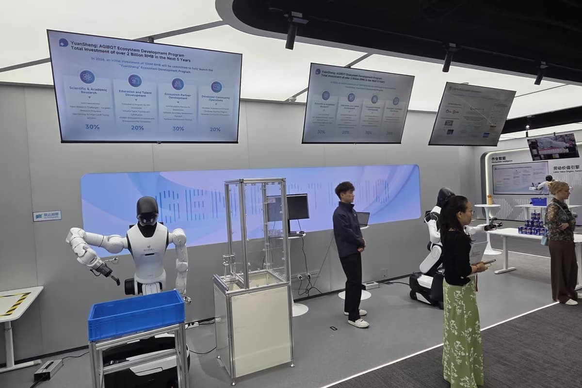 Ein humanoider Roboter greift an einer Arbeitsstation in eine blaue Transportkiste, dahinter Bildschirme und Besucher.