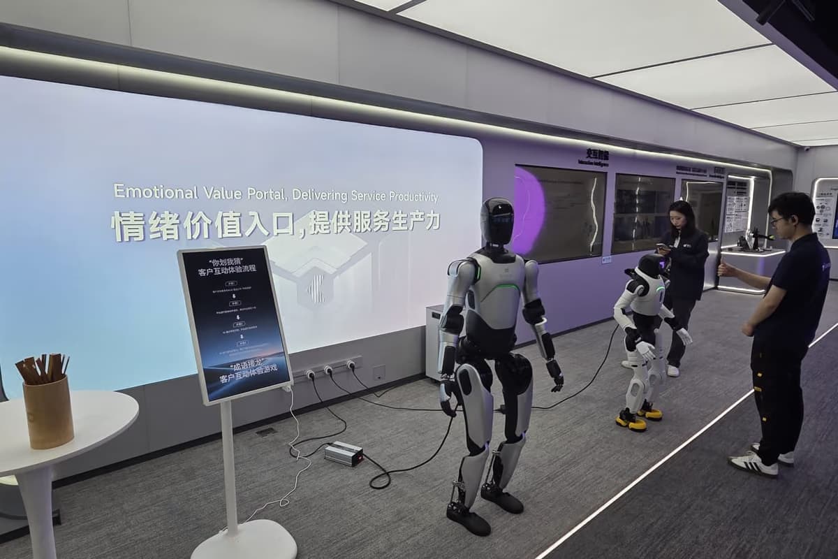 Zwei humanoide Roboter in einem Gang vor einer Bildschirmwand mit chinesischer Schrift, rechts stehen Mitarbeiter.