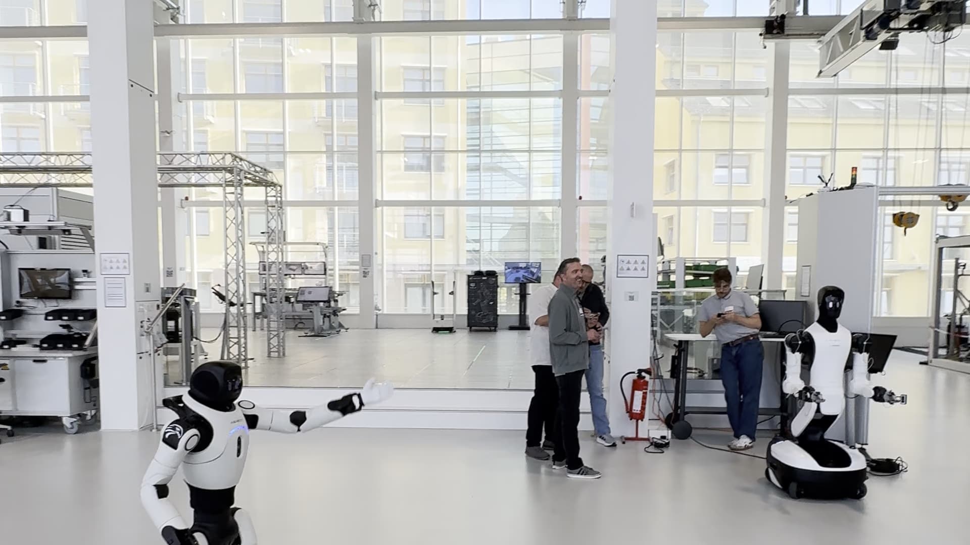 Ein humanoider Roboter geht durch eine helle, hohe Halle mit Fensterfront, im Hintergrund stehen einzelne Personen.