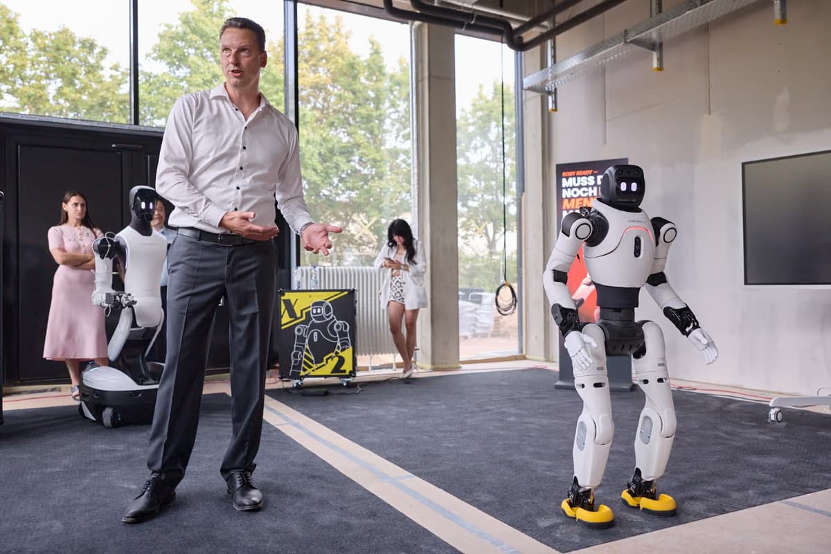 Ein Mann im hellen Hemd spricht mit ausgebreiteten Händen zu Gästen, rechts neben ihm steht ein humanoider Roboter, im Hintergrund ein weiterer.