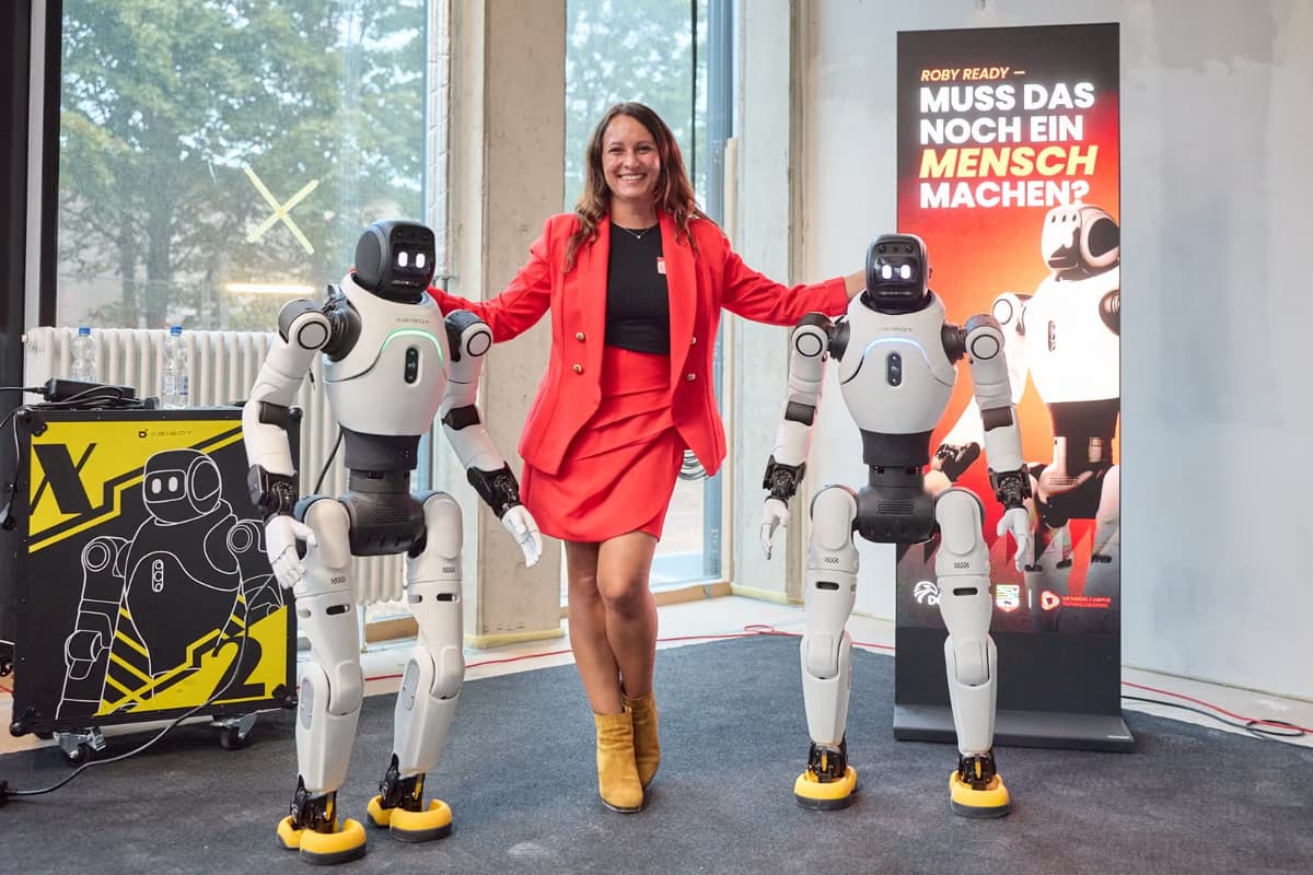 Eine Frau im roten Anzug steht zwischen zwei humanoiden Robotern und legt beiden eine Hand auf die Schulter, rechts ein Aufsteller mit der Frage, ob das noch ein Mensch machen muss.
